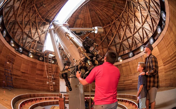 A man holds the controls of a large telescope, pointed toward the sky, as a father and son look on