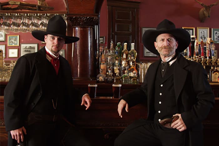 Two men in formal black, Old West clothing stand near a wooden bar