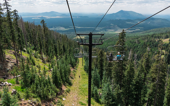View of a chairlift as it descends into a grove of trees with a panoramic view of the mountains behind